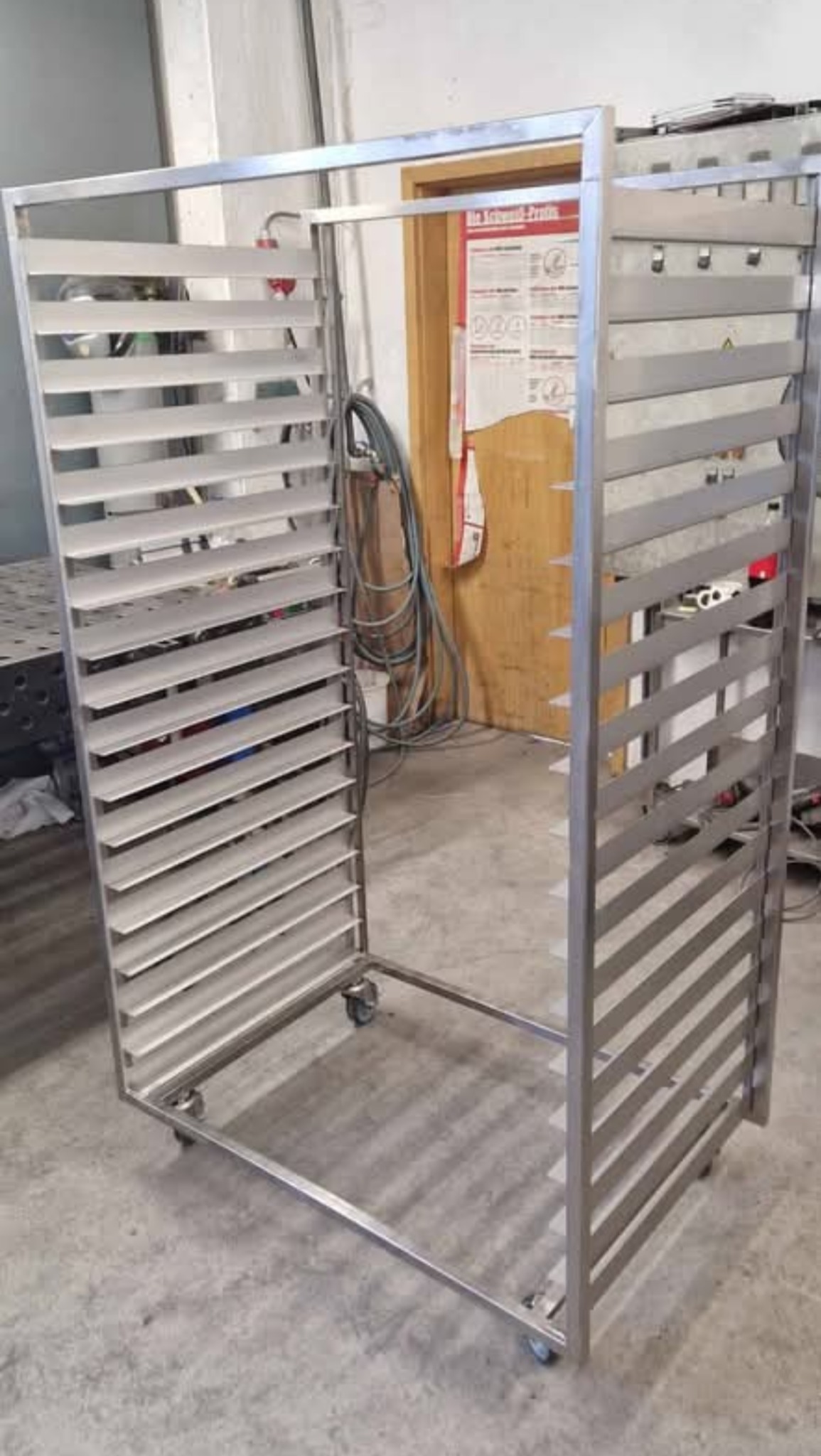 Wózek transportowy ze stali nierdzewnej z półkami szczelinowymi na kółkach, widok z przodu, w tle ściana i drzwi z instrukcją BHP.