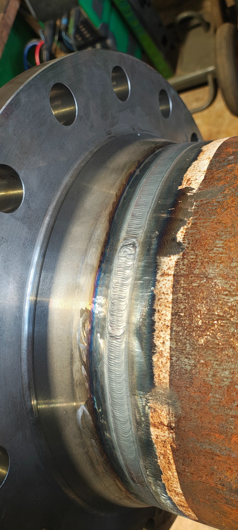 Spoina łącząca rdzewiejącą rurę z kołnierzem, widoczne ślady spawania i opalizujące przebarwienia na metalu. Kołnierz ma otwory na śruby.