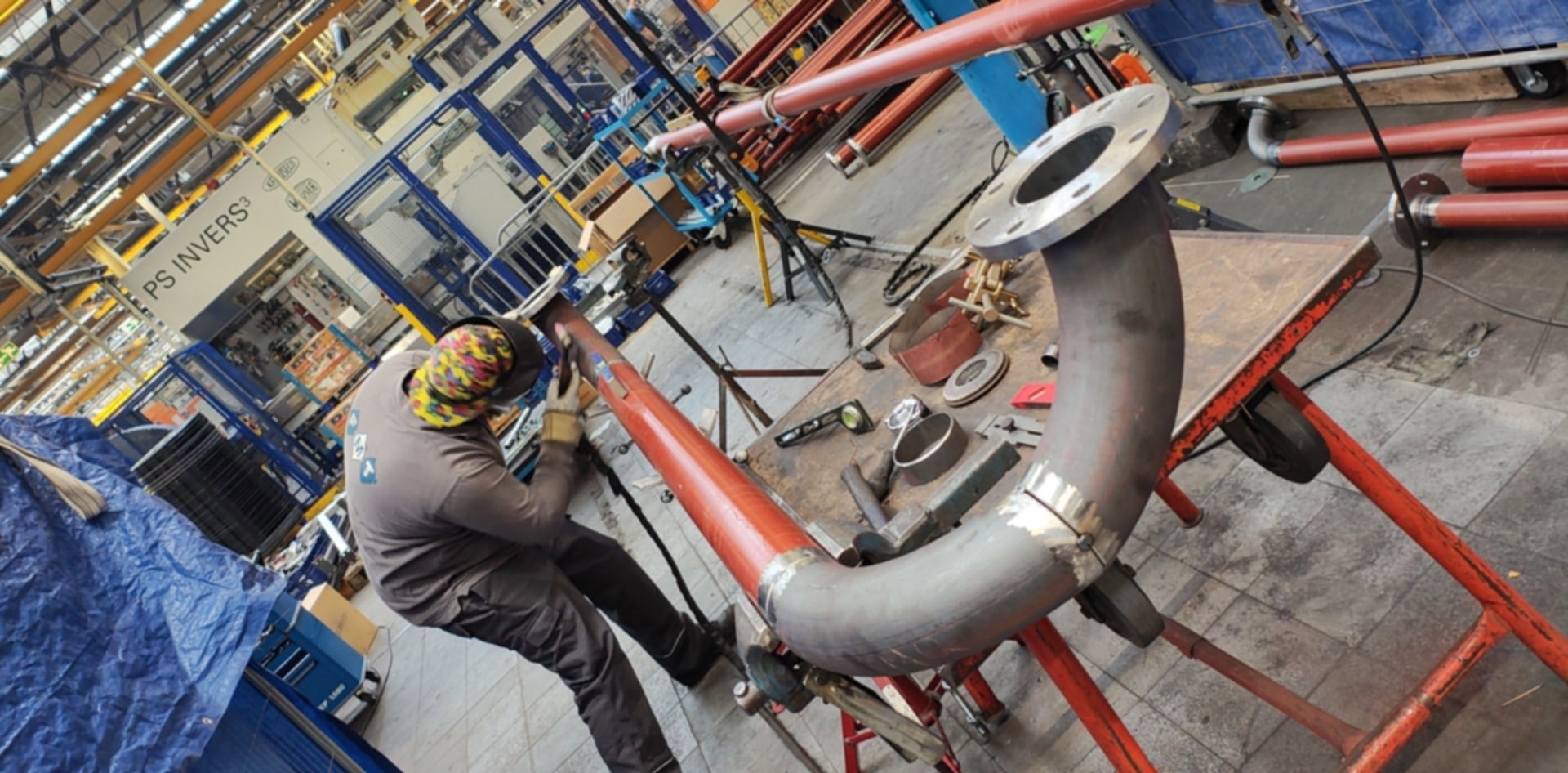 Spawacz w trakcie pracy nad metalową rurą z kołnierzem na warsztacie, widoczne narzędzia i wyposażenie spawalnicze. Praca w hali przemysłowej.