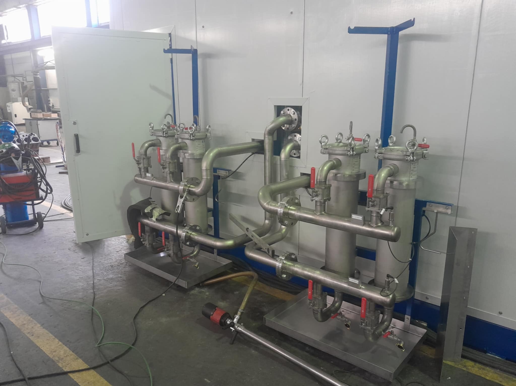 Złożony system filtracji przemysłowej ze stali nierdzewnej, widoczne rury, zawory i zbiorniki filtracyjne. Tło stanowi biała ściana hali produkcyjnej.