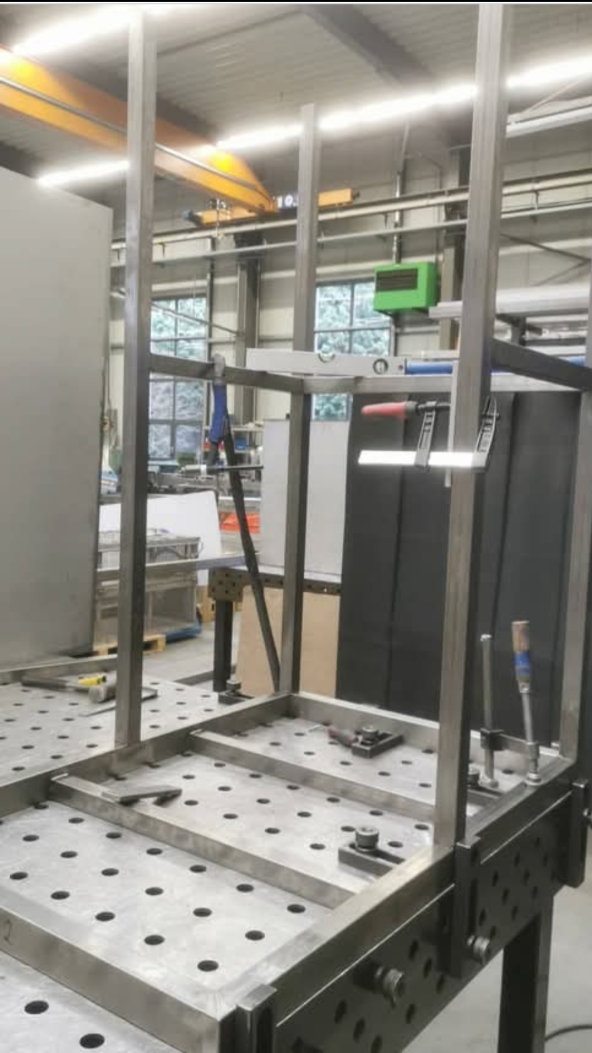 Metalowa konstrukcja w trakcie budowy, spawana na stole z otworami. Widoczne narzędzia i poziomica, w tle hala produkcyjna. Elementy konstrukcji zabezpieczone klamrami.