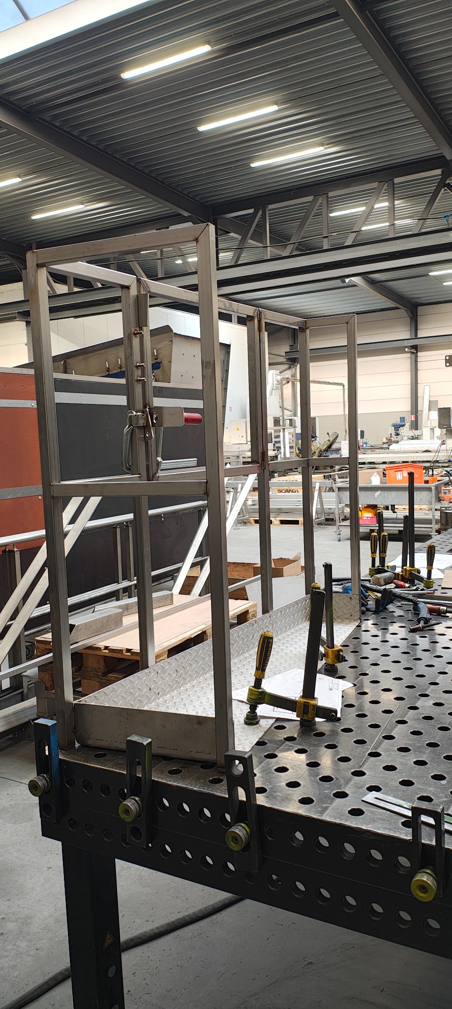 Metalowa konstrukcja z użyciem klamer na perforowanym stole spawalniczym w hali produkcyjnej. Elementy mocowane do spawania. Widoczne narzędzia i akcesoria.