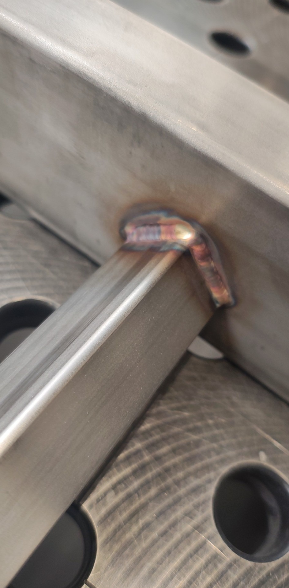 Metalowa konstrukcja spawana, widoczny estetyczny spaw z opalizującymi przebarwieniami termicznymi. Połączenie kątowe na stole spawalniczym.