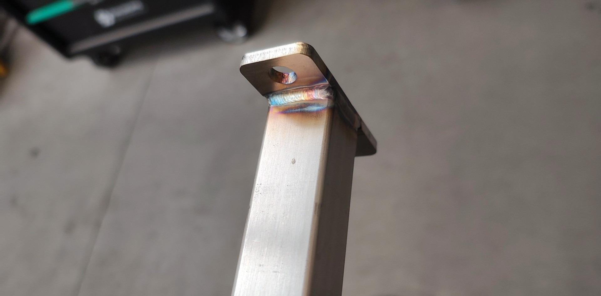 Szlifowany profil stalowy z przyspawaną płytką z otworem. Widoczne ślady spawania TIG z charakterystycznymi kolorami. Detal konstrukcyjny.