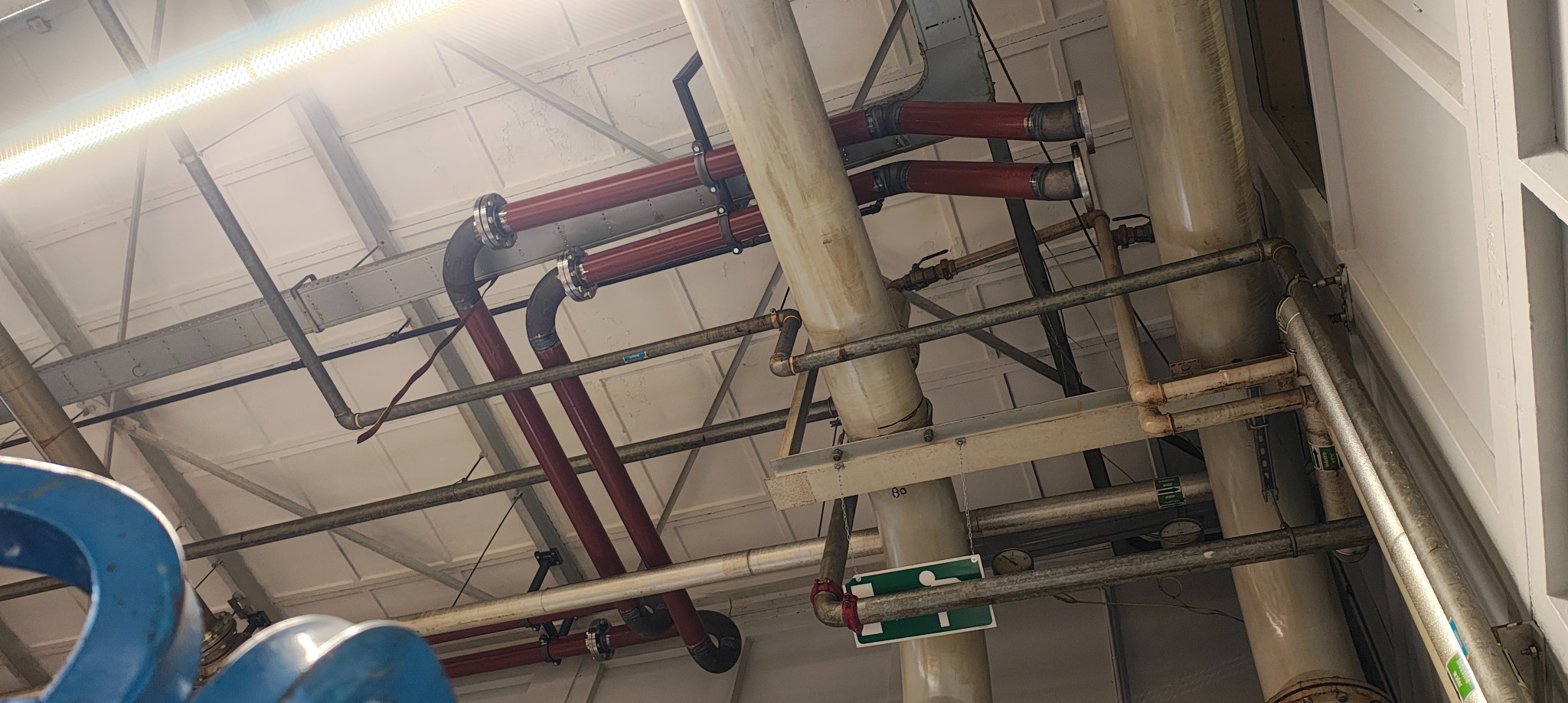 Złożona instalacja przemysłowa z rurami w kolorze czerwonym i szarym, zawieszona pod sufitem. Widoczne elementy konstrukcyjne budynku i oznakowanie bezpieczeństwa.