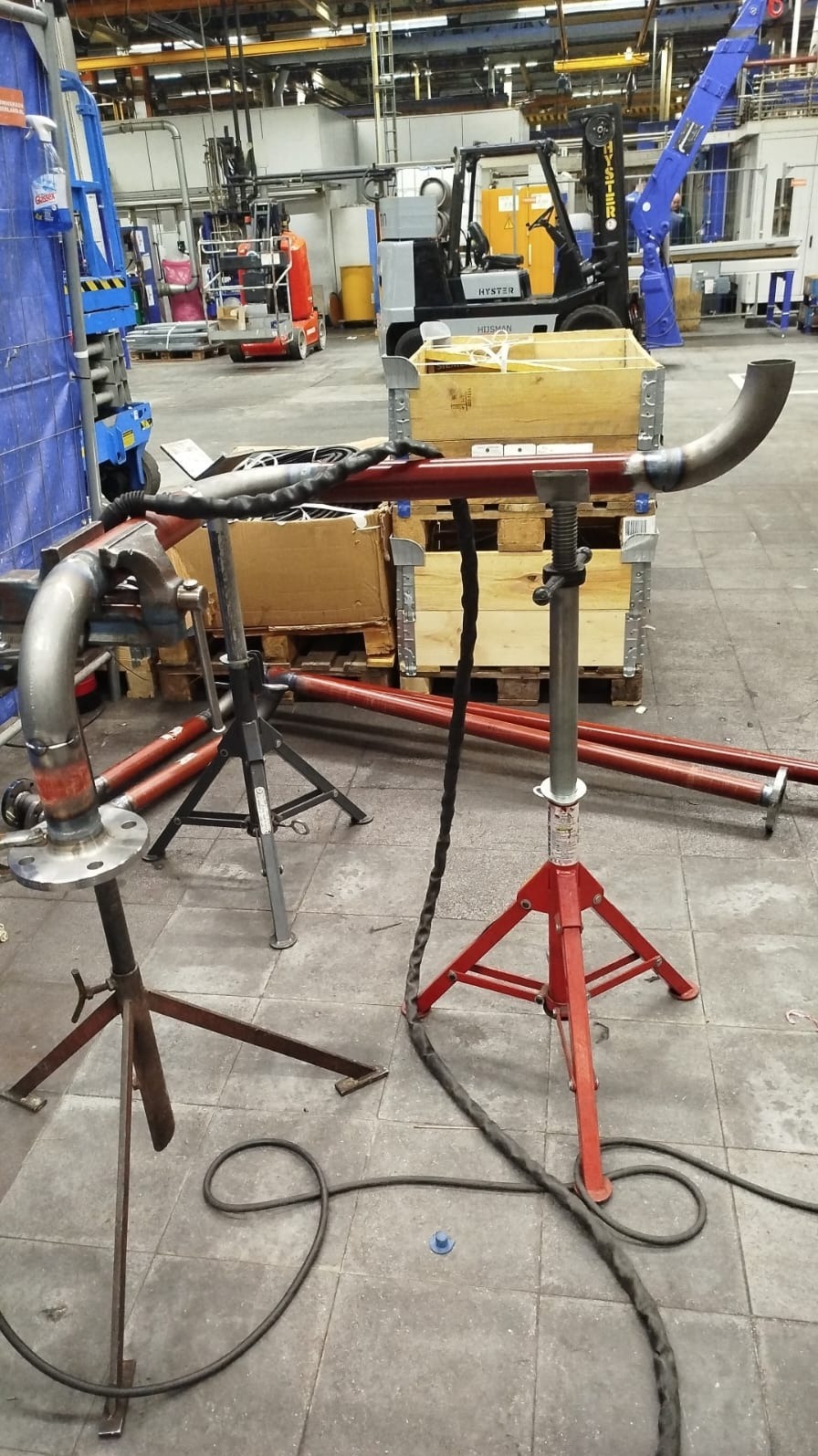 Spawane rury na stojakach w warsztacie, widoczny spaw, palety, wózek widłowy w tle. Przewód spawalniczy leży na podłodze. Szlifowane spawy na rurach.