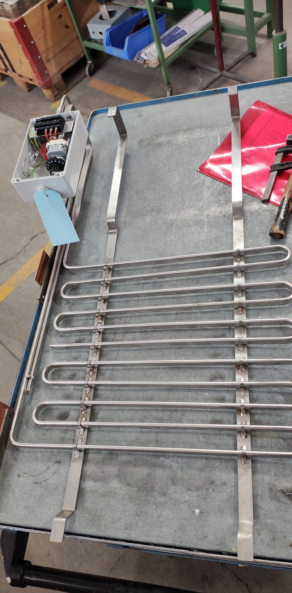 Metalowa konstrukcja z rur w kształcie litery U, połączona płaskownikami, leży na metalowym stole. Widoczna skrzynka elektryczna z podłączonymi kablami.