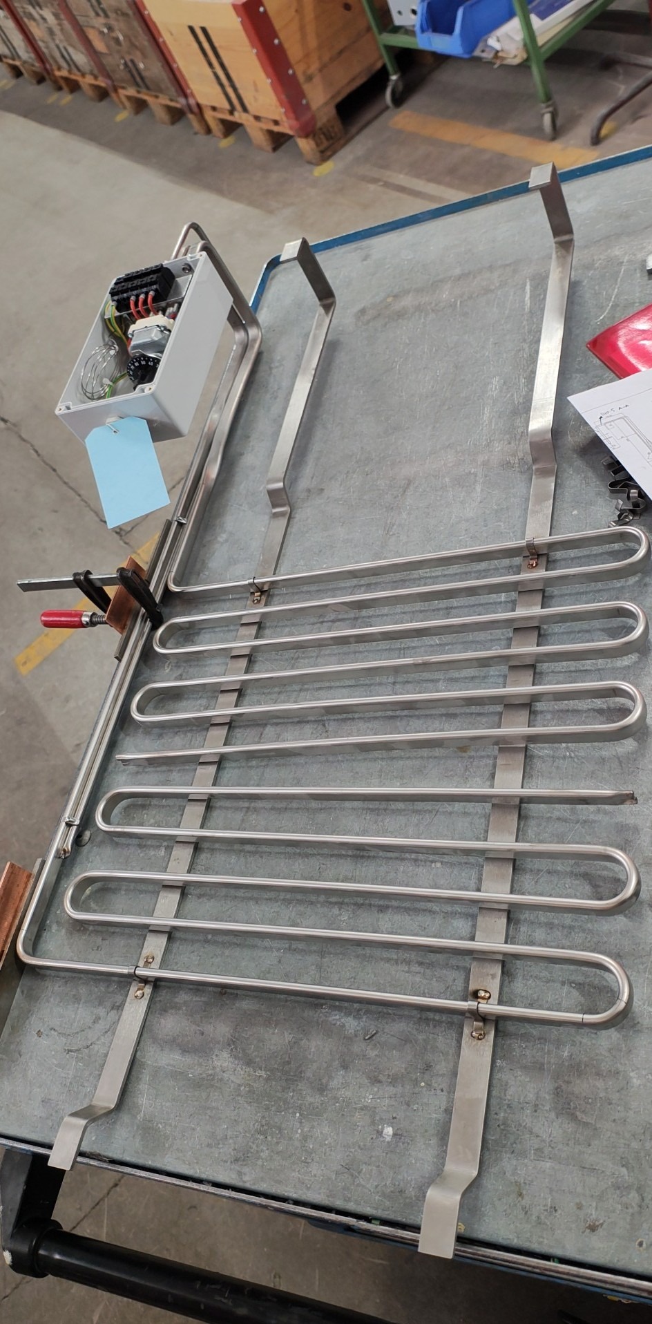 Metalowa konstrukcja z rurek, prawdopodobnie element grzejny, na szarym stole warsztatowym. Widoczna skrzynka elektryczna i zacisk stolarski.