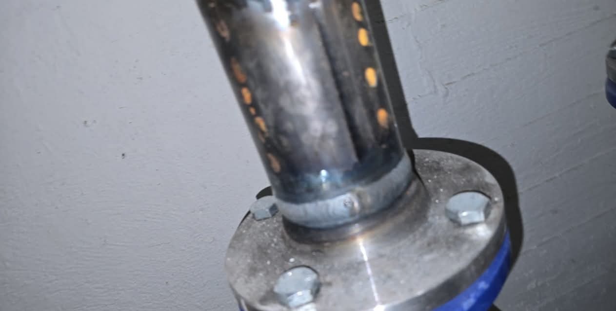 Detal spawu łączącego rurę ze stalową podstawą, widoczne ślady spawania i śruby mocujące. Szara ściana w tle, metaliczna powierzchnia z refleksami.