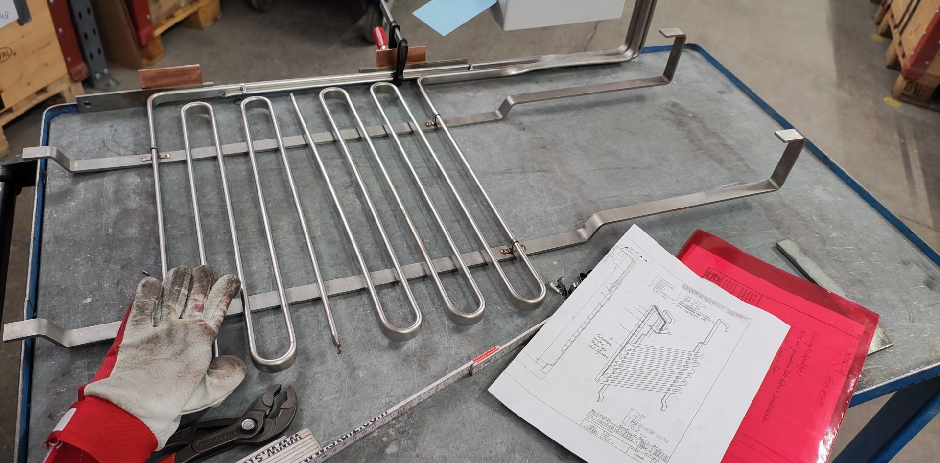 Konstrukcja metalowa z rurek i płaskowników na stole warsztatowym, obok rękawica robocza, narzędzia i rysunek techniczny. Widoczne ślady spawania.