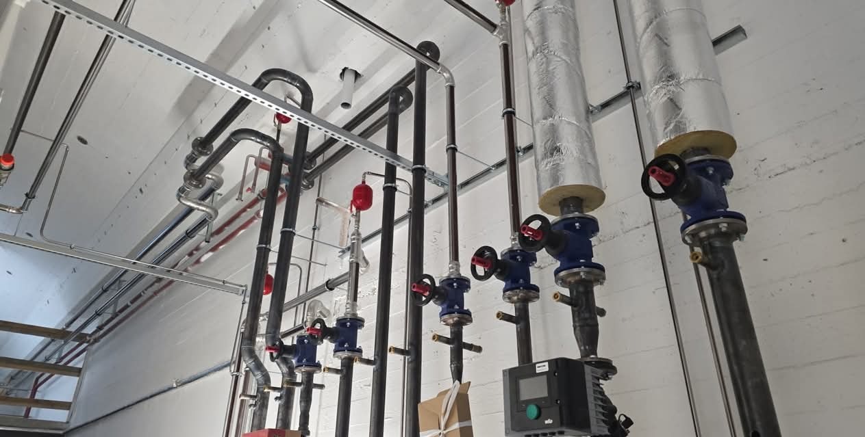 Instalacja przemysłowa z rurami, zaworami i izolacją, zamontowana przy białej, teksturowanej ścianie. Widoczne fragmenty konstrukcji wsporczej.