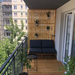 Stolar-RS - Przytulny balkon w bloku z drewnianą podłogą i ażurową ścianką. Sofa z poduszkami, stolik i wiszące doniczki tworzą relaksującą przestrzeń miejską.