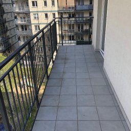 Stolar-RS - Nowoczesny balkon z szarą terakotą, metalową balustradą i siatką zabezpieczającą. Widok na osiedle mieszkaniowe zieleń. Jasne, naturalne oświetlenie.