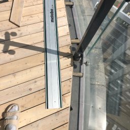 Stolar-RS - Montaż drewnianego tarasu z balustradą szklaną. Widoczna szyna tnąca Metabo, deski tarasowe, mocowania balustrady i odbicie otoczenia w szkle.