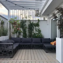 Stylowa aranżacja tarasu z drewnianą podłogą i pergolą oświetloną girlandą. Wygodna, grafitowa sofa z poduszkami i rośliny w donicach tworzą przytulny klimat.