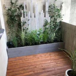 Drewniany taras z roślinami w donicach i panelami dekoracyjnymi na ścianie. Jasne drewno, ciepłe brązowe deski tarasowe. Oświetlenie punktowe na ścianie.