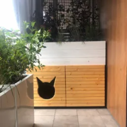 Balkon z drewnianą zabudową, donicami z zielenią i wyciętym otworem w kształcie kociej głowy w dolnej części. Ściana z drewnianą okładziną.