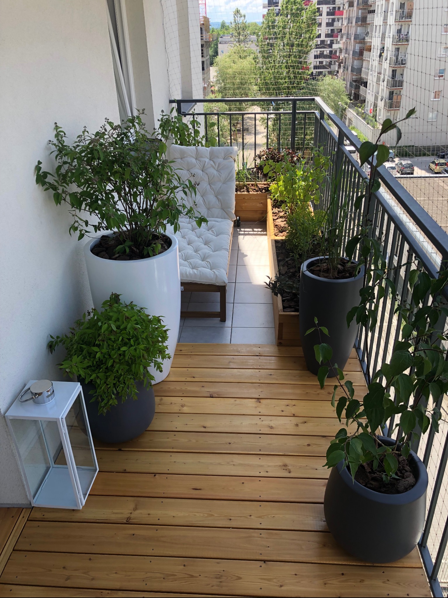 Jasny balkon z drewnianą podłogą, roślinami w donicach i metalową balustradą. W tle widoczne budynki mieszkalne i siatka zabezpieczająca. Widok z góry.