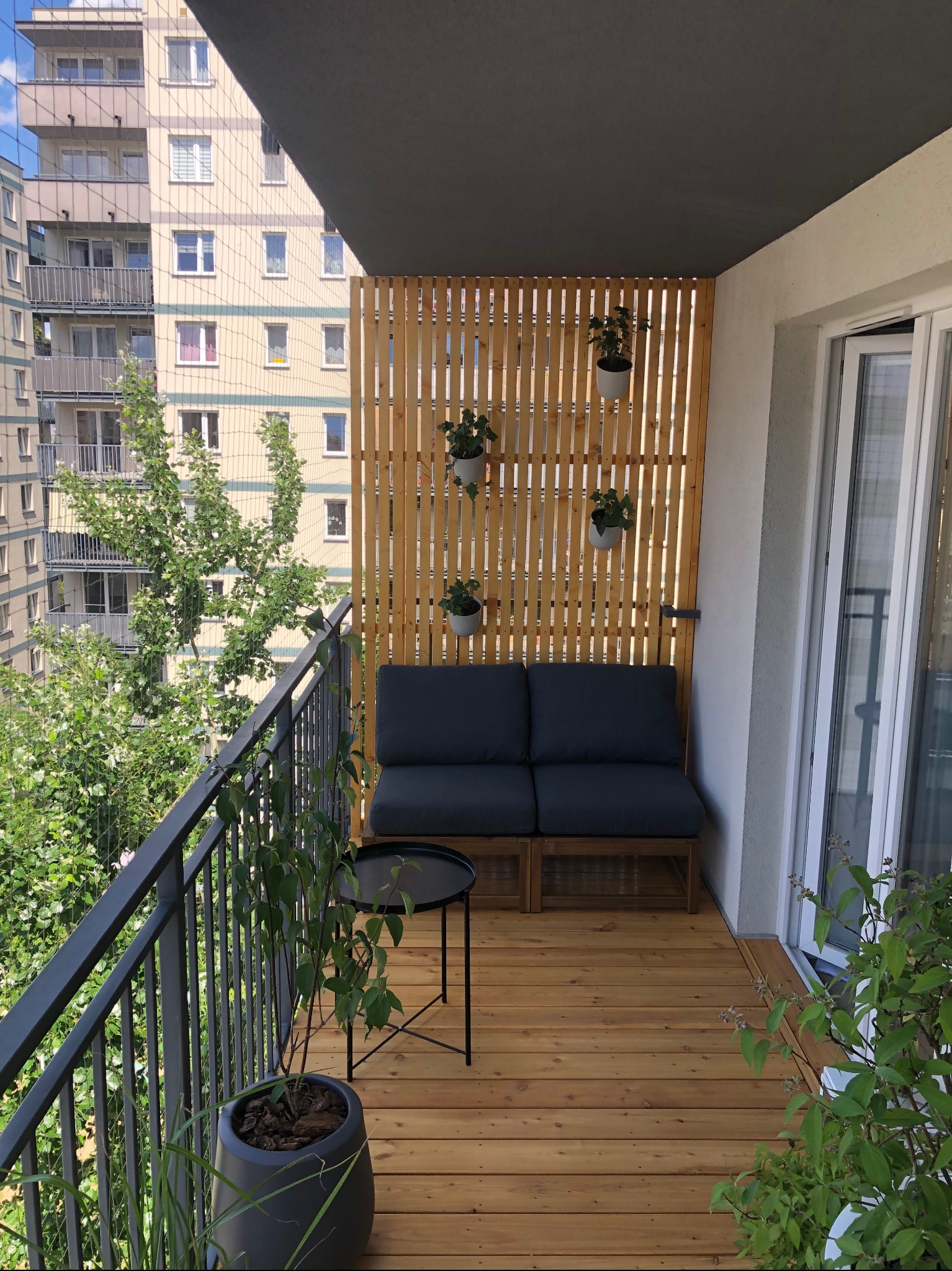 Przytulny balkon w bloku z drewnianą podłogą i ażurową ścianką. Sofa z poduszkami, stolik i wiszące doniczki tworzą relaksującą przestrzeń miejską.