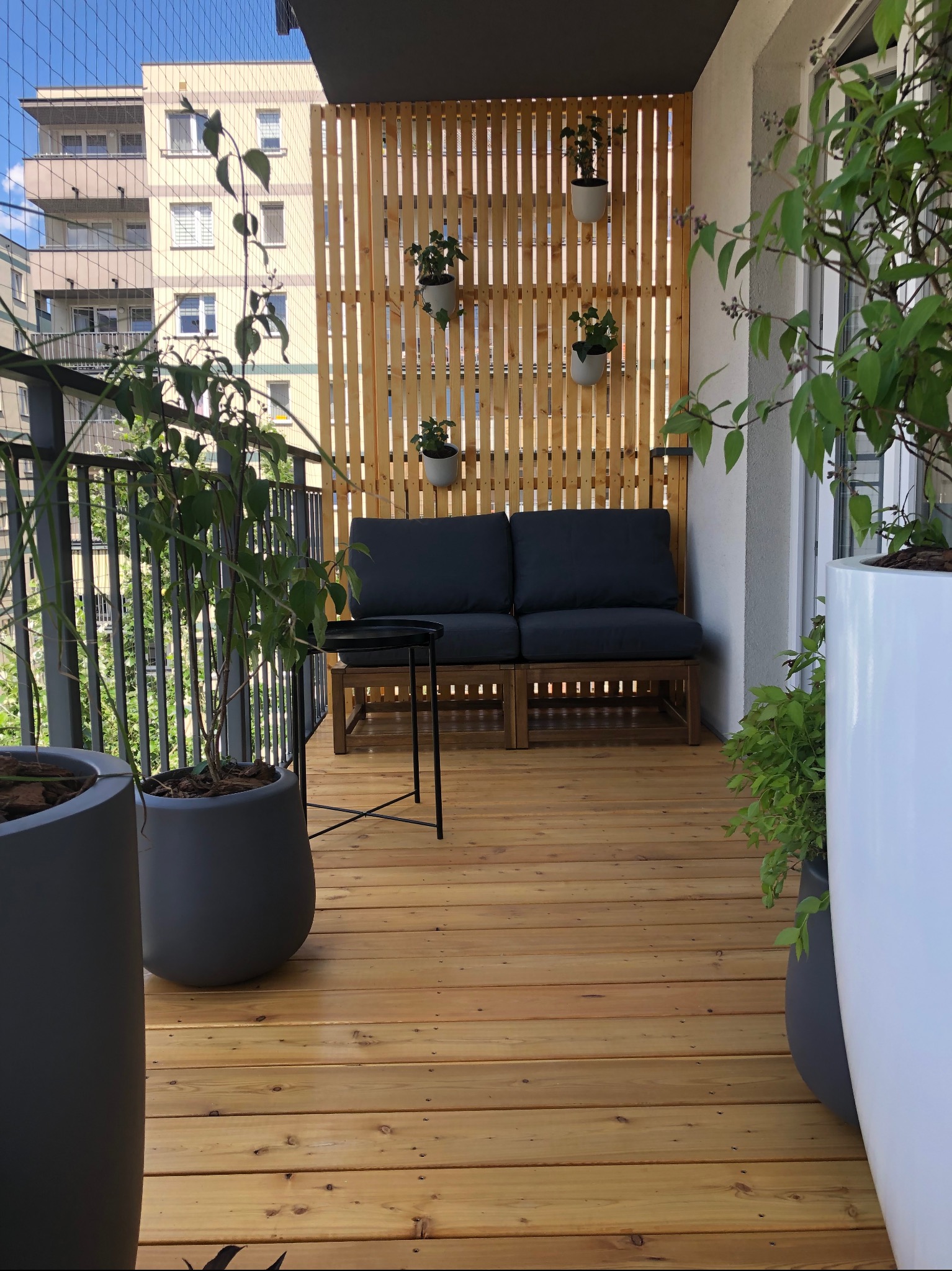 Jasny, drewniany balkon z drewnianą, ażurową ścianką, sofą z szarymi poduchami, stolikiem i roślinami w donicach. Widok na budynek mieszkalny zza balustrady.