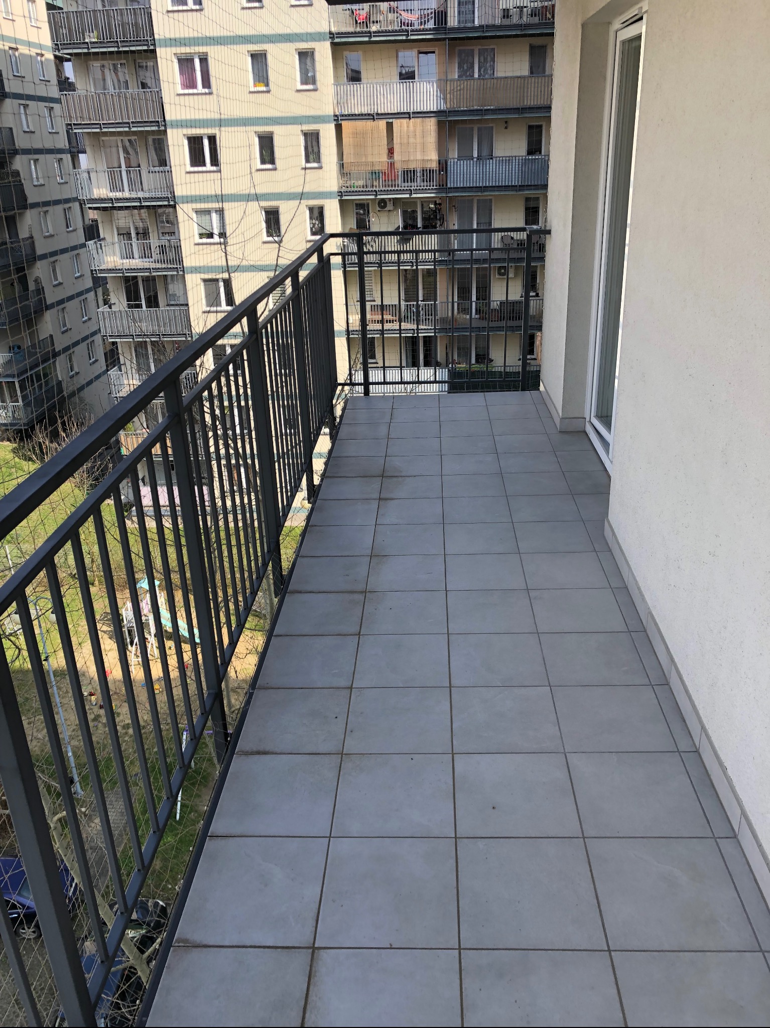 Nowoczesny balkon z szarą terakotą, metalową balustradą i siatką zabezpieczającą. Widok na osiedle mieszkaniowe zieleń. Jasne, naturalne oświetlenie.