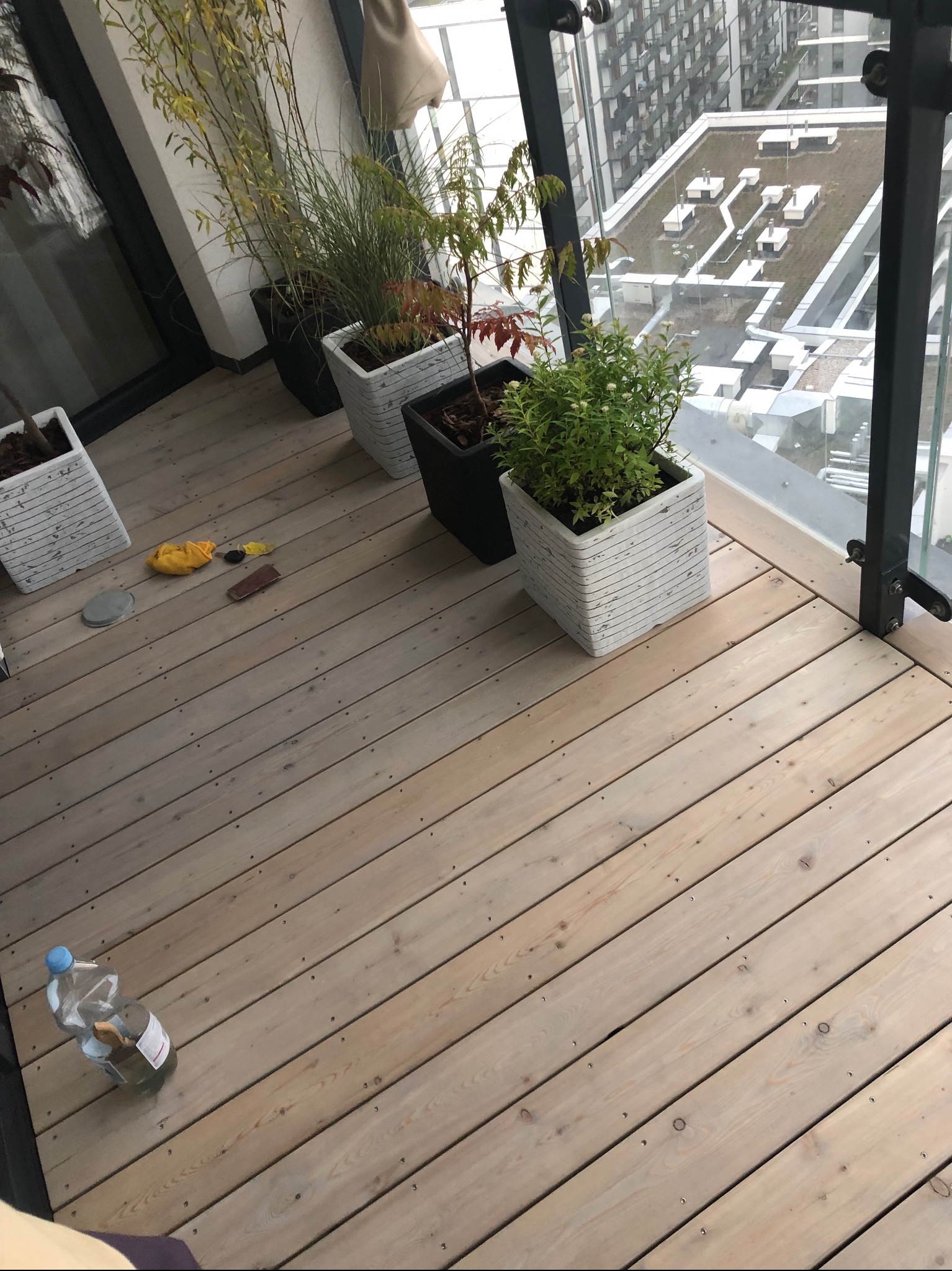 Jasny drewniany taras na balkonie z roślinami w donicach i szklaną balustradą, widok na budynki w tle. Widać ślady pracy: butelka wody, szmatka i gąbka.
