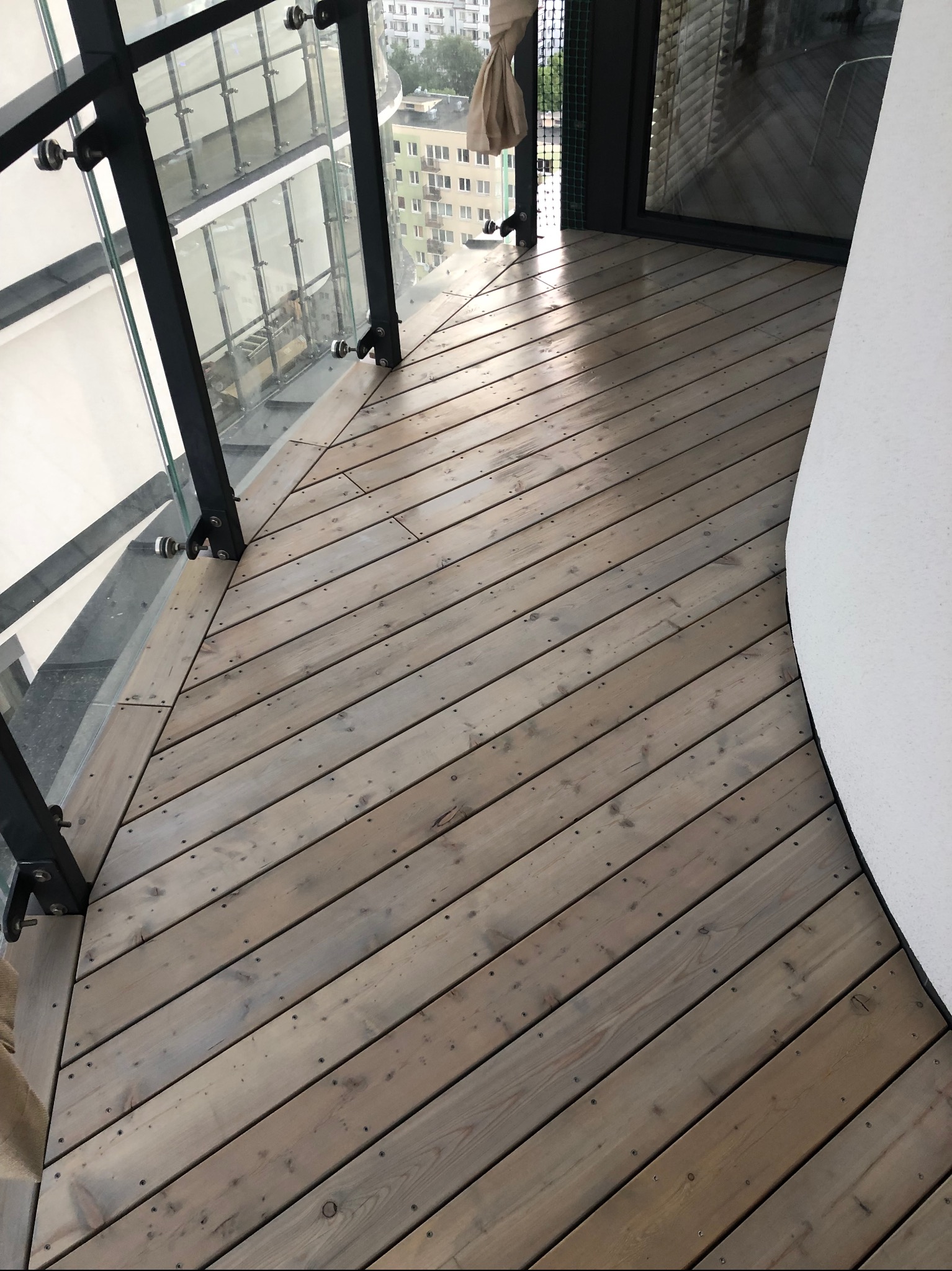 Drewniany taras na balkonie z szklaną balustradą i widokiem na miasto. Deski ułożone ukośnie, widoczne mocowania balustrady. Nowoczesne wykończenie.