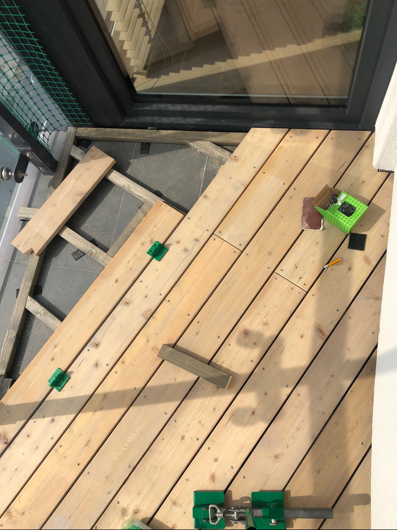 Montaż desek tarasowych na balkonie, widoczne narzędzia i elementy konstrukcji. Deski ułożone pod kątem, drewno jasne, w tle okno. Prace w toku.