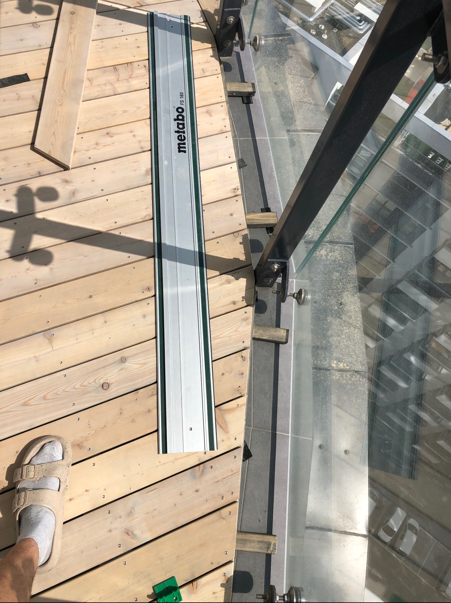 Montaż drewnianego tarasu z balustradą szklaną. Widoczna szyna tnąca Metabo, deski tarasowe, mocowania balustrady i odbicie otoczenia w szkle.