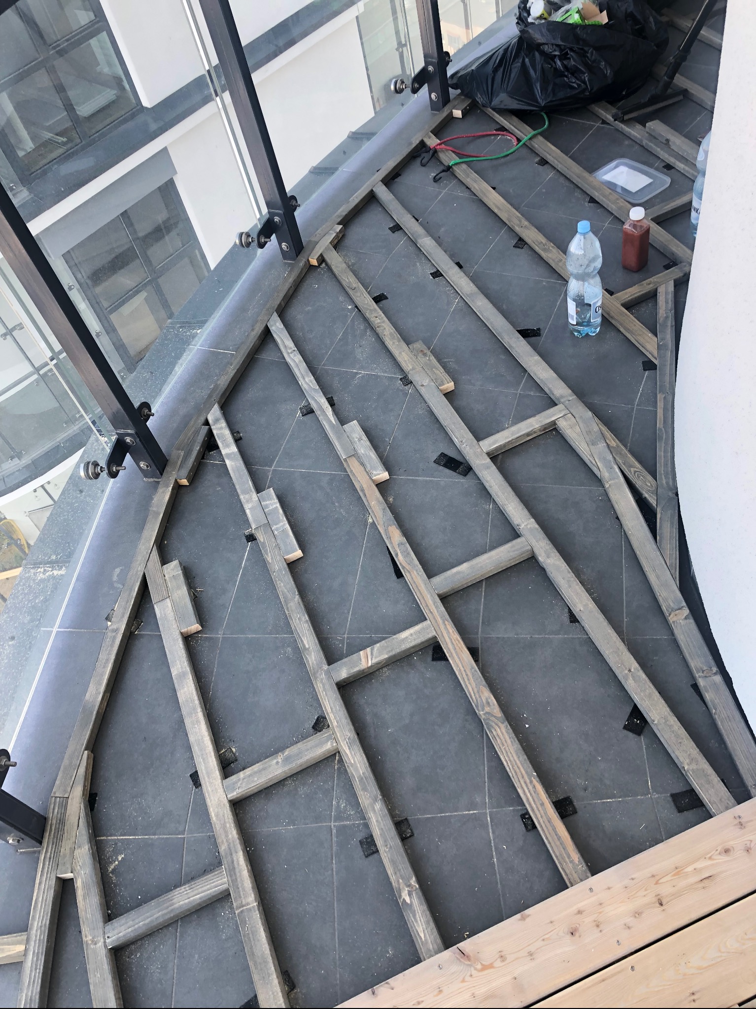 Konstrukcja pod taras drewniany w Brzeźnicy, widok z góry. Szare deski ułożone na ciemnych płytkach, przygotowane do montażu. Elementy szklanej balustrady.