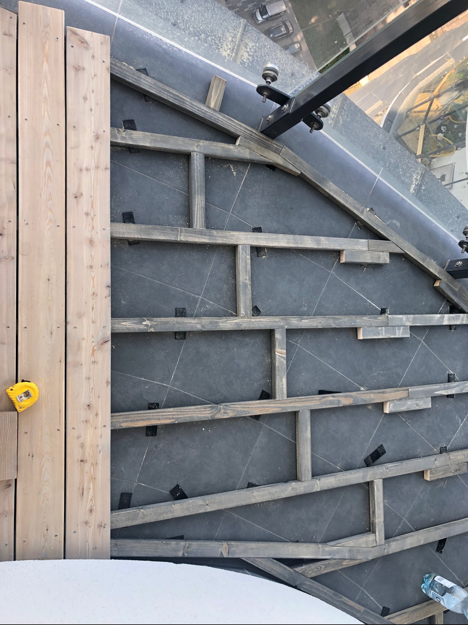 Montaż drewnianego tarasu na balkonie. Widoczny szkielet konstrukcji, deski i miara. Kąt z góry, częściowo widoczny widok z balkonu.