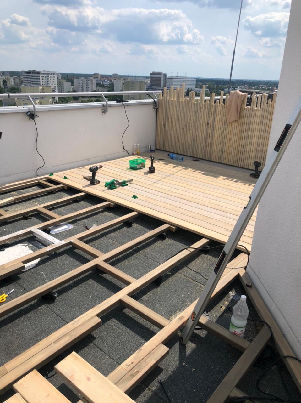 Montaż jasnego tarasu na dachu budynku. Częściowo ułożone deski, widoczny stelaż i narzędzia. W tle zabudowa z pionowych desek i panorama miasta.