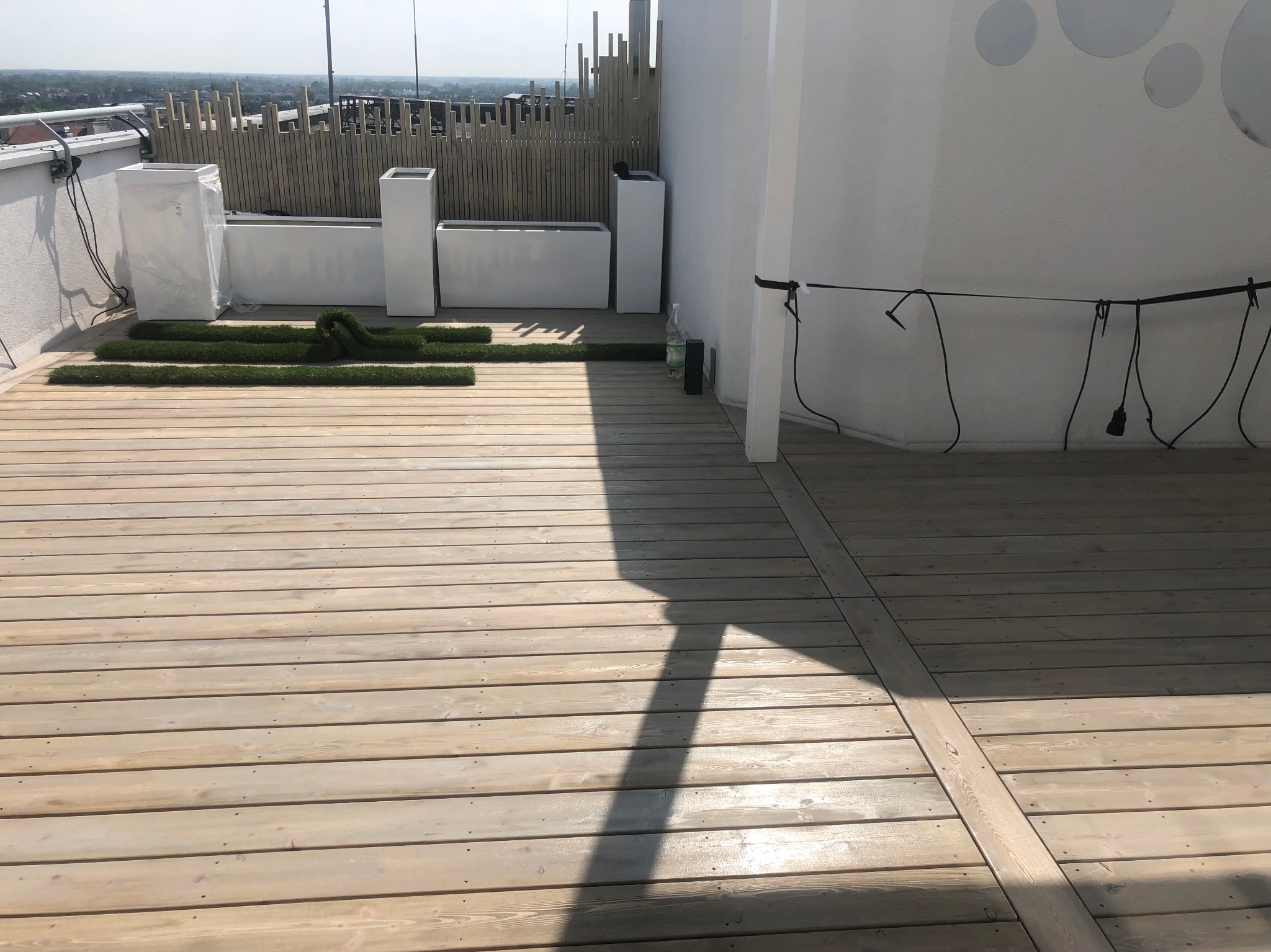 Jasny taras z desek na dachu budynku, widok na miasto w tle. Donice i fragmenty sztucznej trawy dodają zieleni. Minimalistyczny design.