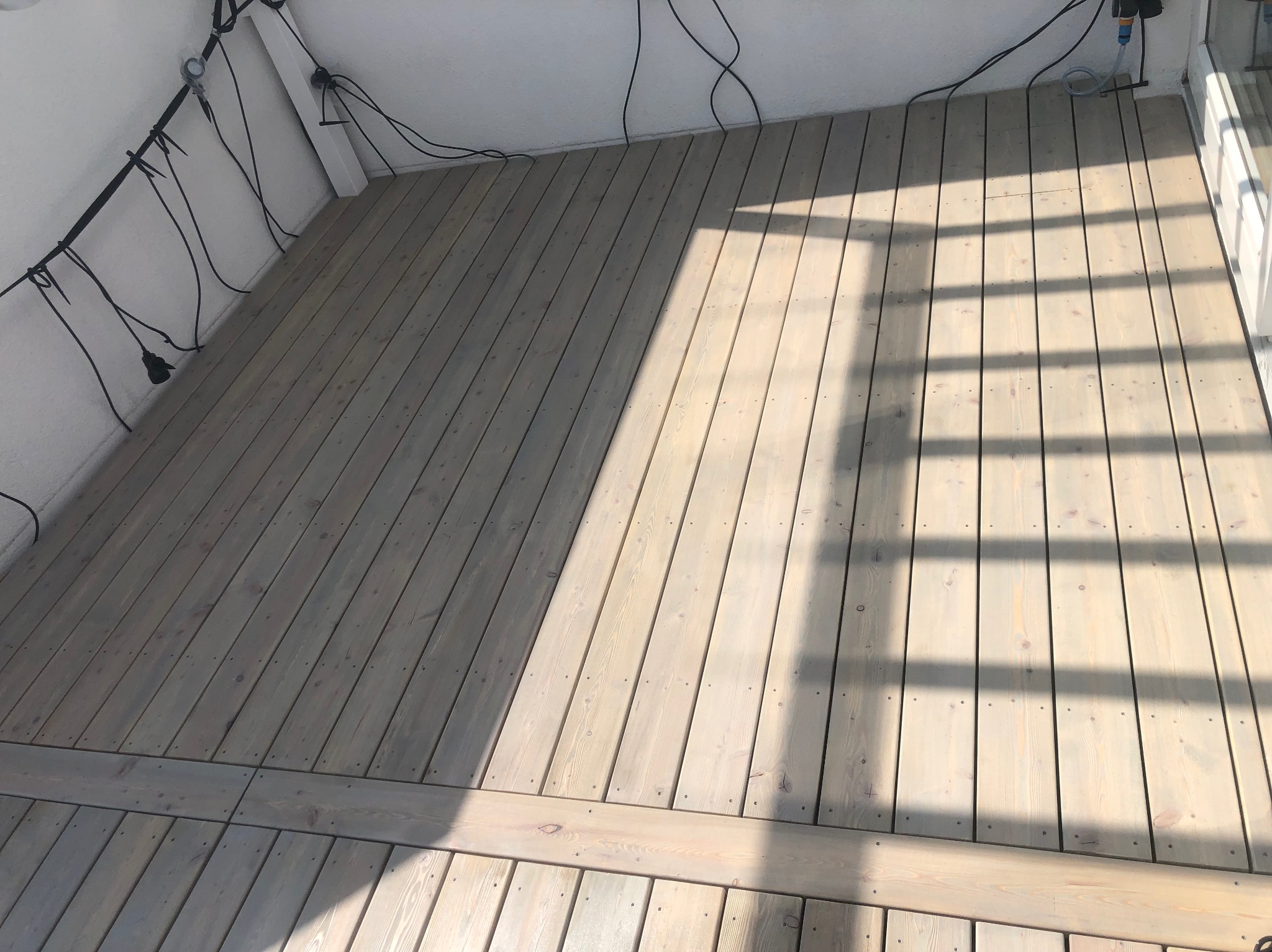 Jasny, pomalowany na szaro drewniany taras z widocznymi śladami światła słonecznego. Deski ułożone równolegle, z widocznymi mocowaniami. W tle biała ściana i kable.