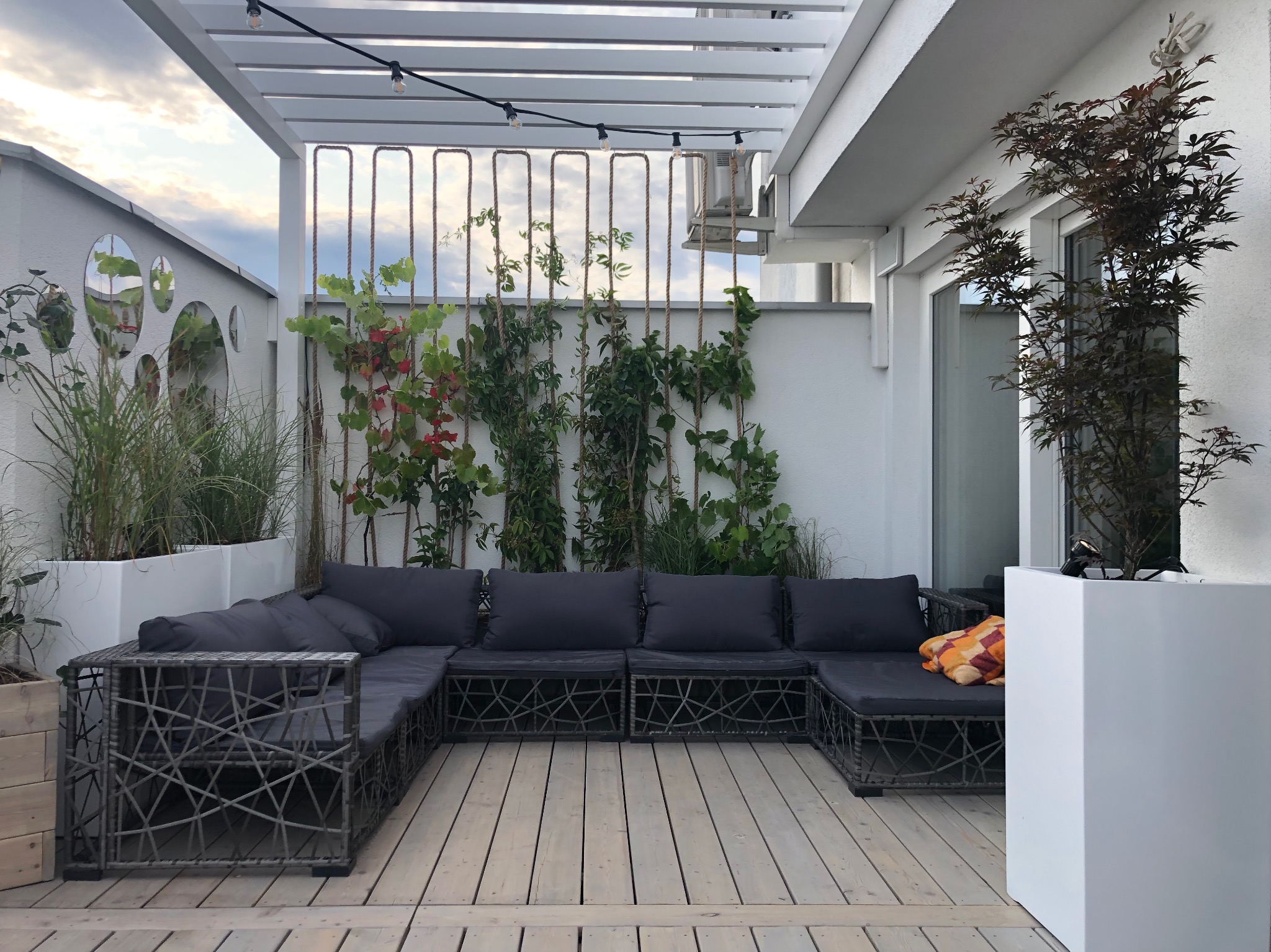 Stylowa aranżacja tarasu z drewnianą podłogą i pergolą oświetloną girlandą. Wygodna, grafitowa sofa z poduszkami i rośliny w donicach tworzą przytulny klimat.