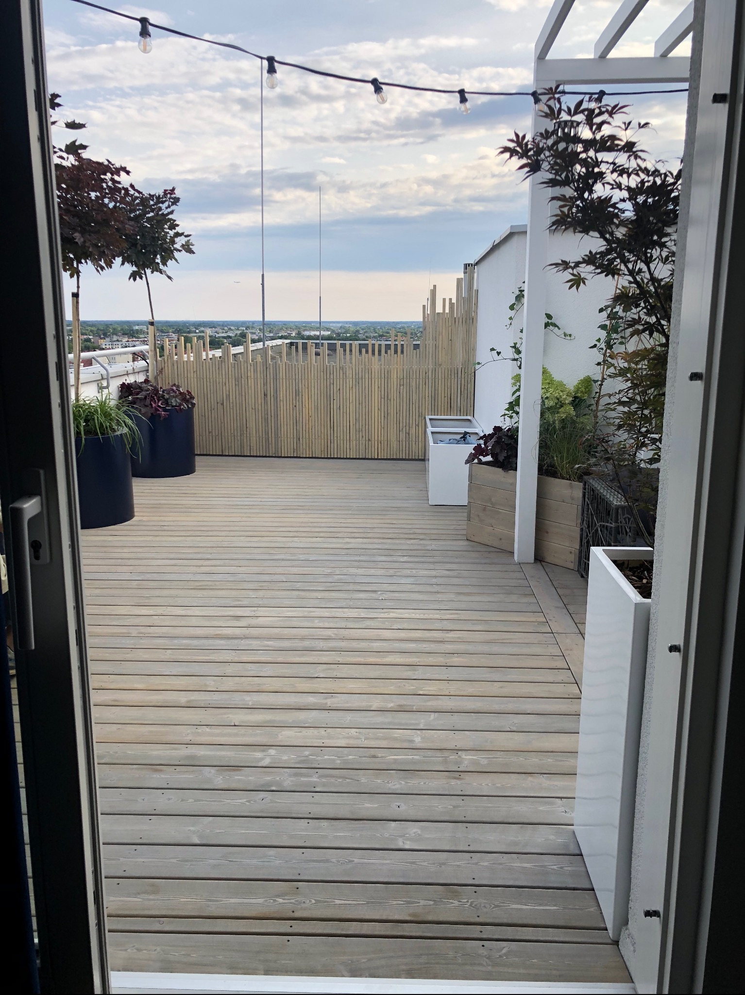 Drewniany taras na dachu budynku z ażurowym ogrodzeniem, donicami i oświetleniem, widok z wnętrza przez otwarte drzwi.