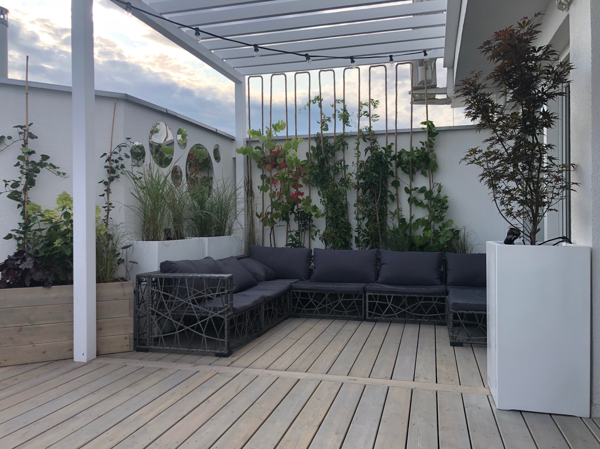 Jasny taras z drewnianą podłogą i szarą sofą z poduszkami. Na ścianie dekoracyjne lustra i pnącza. Nad sofą pergola z oświetleniem. Rośliny w donicach.