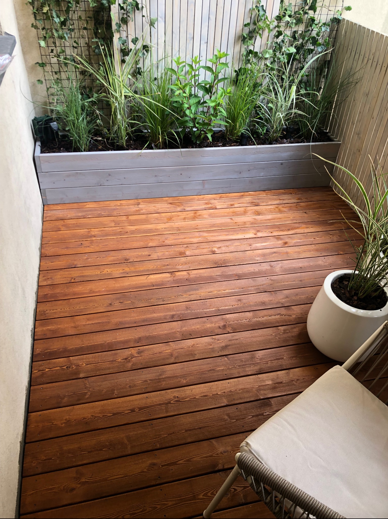 Drewniany taras z jasną, pionową zabudową i roślinami w donicy. Ujęcie z góry, widoczny fragment krzesła.