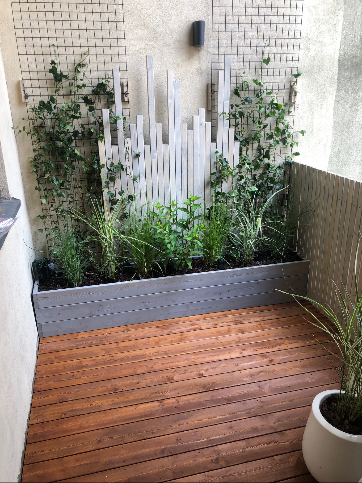 Drewniany taras z roślinami w donicach i panelami dekoracyjnymi na ścianie. Jasne drewno, ciepłe brązowe deski tarasowe. Oświetlenie punktowe na ścianie.