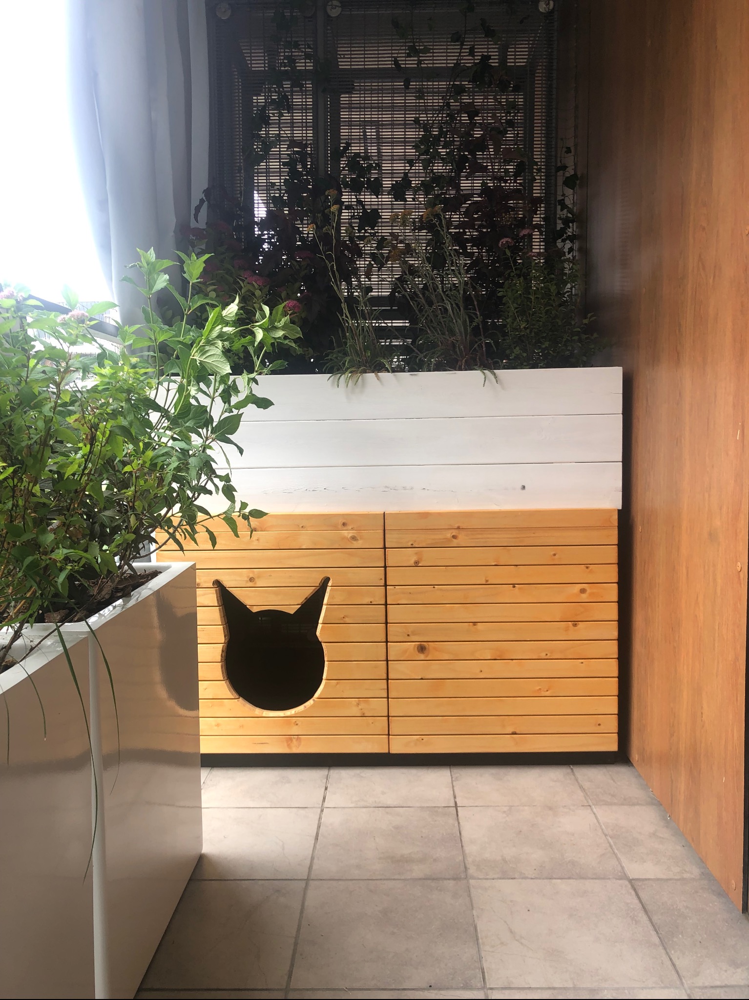 Balkon z drewnianą zabudową, donicami z zielenią i wyciętym otworem w kształcie kociej głowy w dolnej części. Ściana z drewnianą okładziną.
