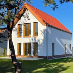 Biały dom z pomarańczową dachówką i oknami, widziany zza drzewa, w słoneczny dzień. Minimalistyczny styl, świeżo wykończony, z zadbanym trawnikiem.