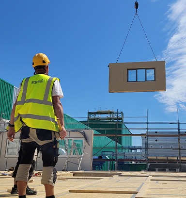 Montaż modułu z oknem na budowie domu modułowego. Pracownik w kamizelce i kasku nadzoruje proces. W tle rusztowania i błękitne niebo.