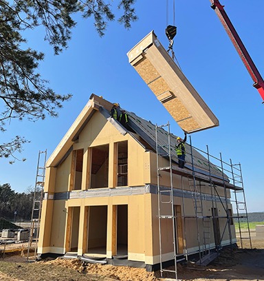 Montaż modułowego domu jednorodzinnego z użyciem dźwigu. Widoczne elementy konstrukcyjne i rusztowania, błękitne niebo w tle.