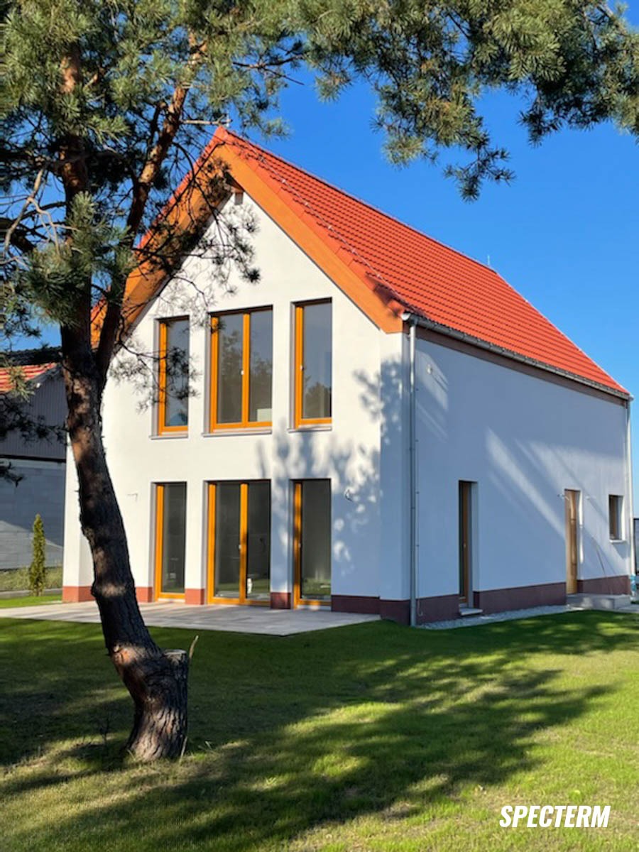 Biały dom z pomarańczową dachówką i oknami, widziany zza drzewa, w słoneczny dzień. Minimalistyczny styl, świeżo wykończony, z zadbanym trawnikiem.