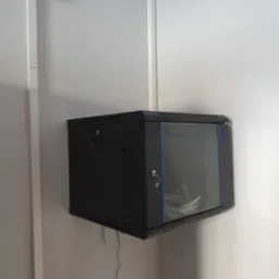 Czarna szafa rack wisząca na ścianie, z widocznymi kablami wewnątrz, zabezpieczona zamkami. Widoczne elementy instalacji elektrycznej na ścianie.