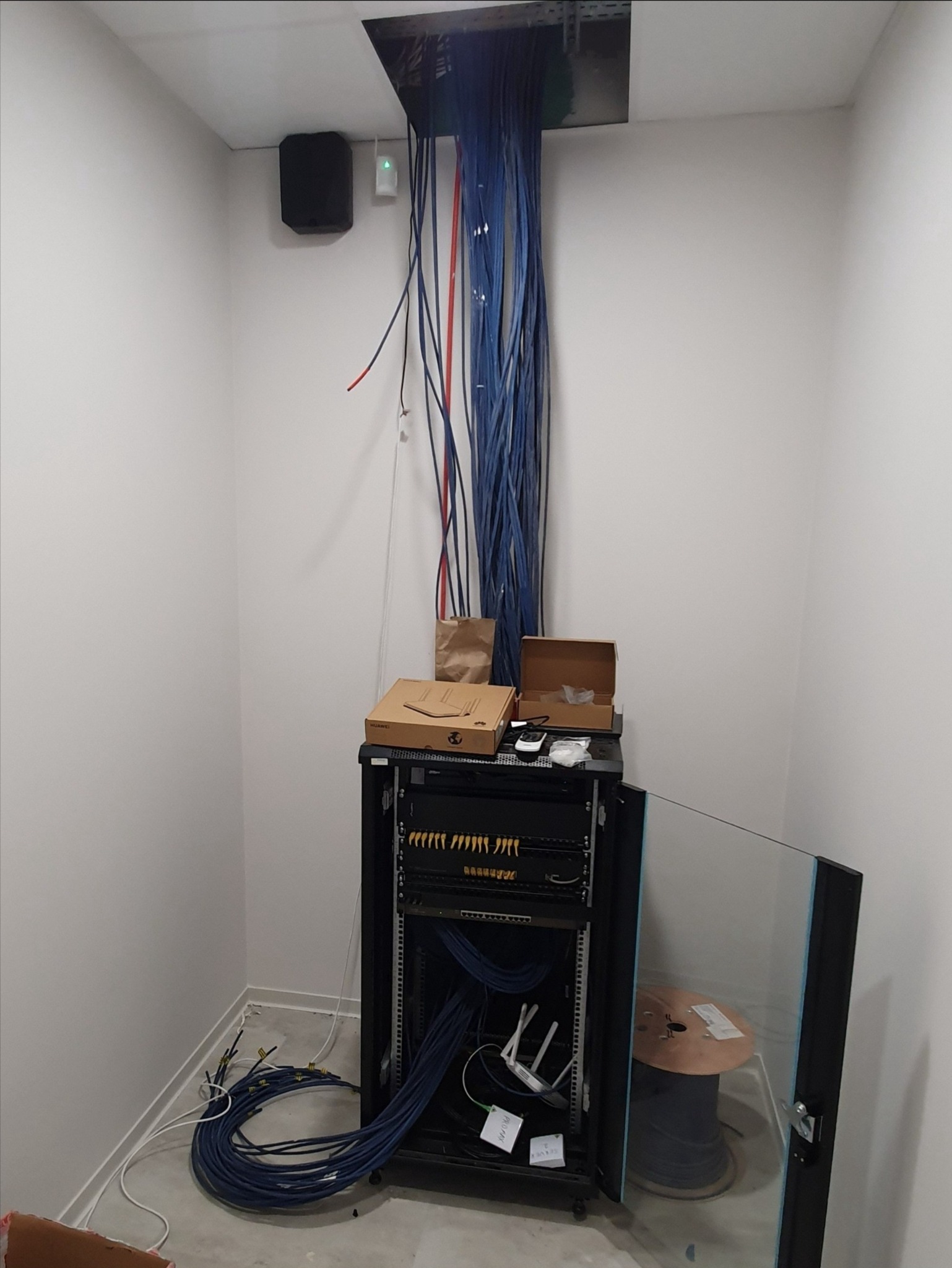 Szafa rack z kablem sieciowym, routerem i akcesoriami w pomieszczeniu z otworem w suficie, z którego wystają przewody. Widoczne pudełka i elementy instalacji.