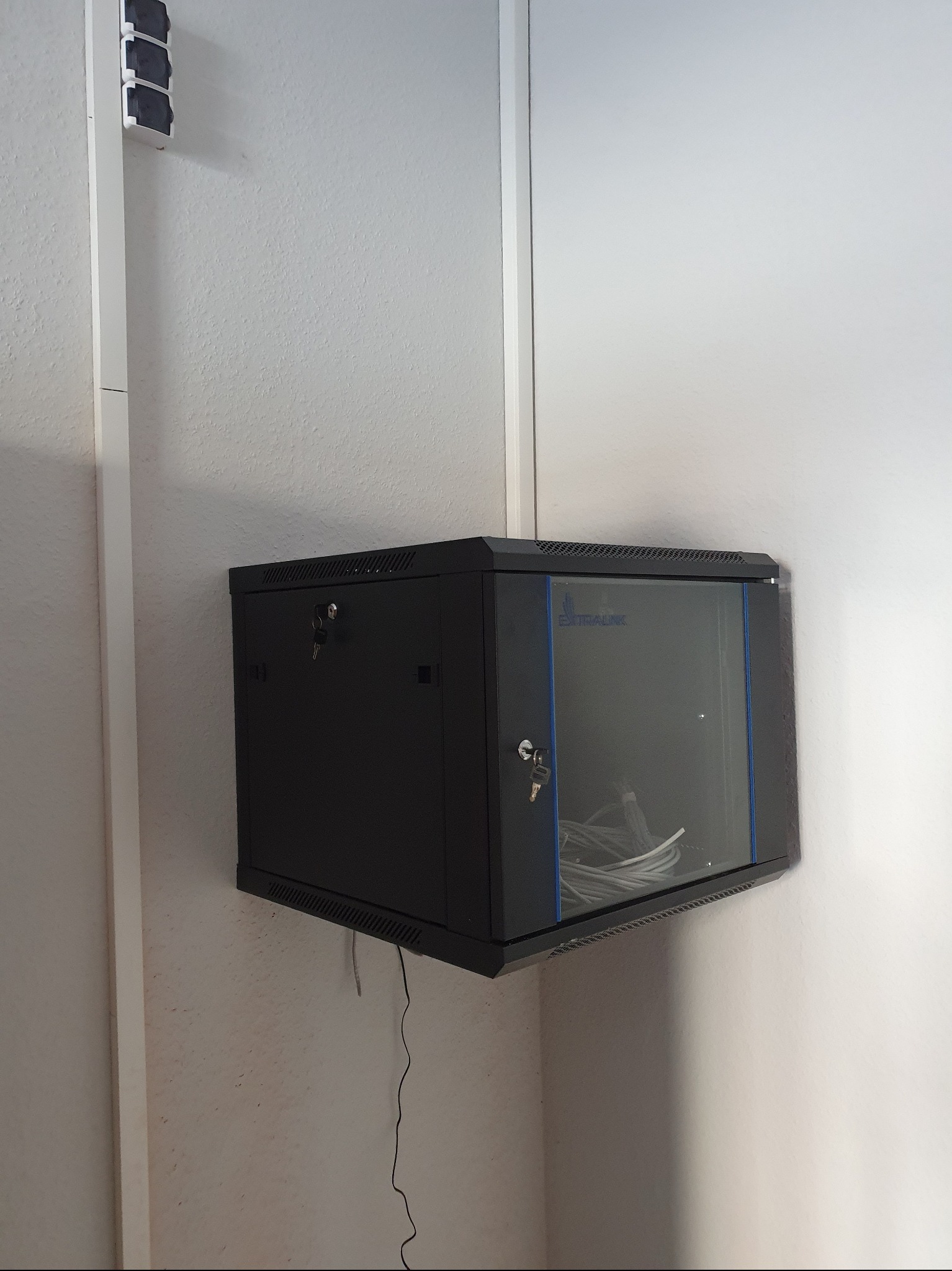 Czarna szafa rack wisząca na ścianie, z widocznymi kablami wewnątrz, zabezpieczona zamkami. Widoczne elementy instalacji elektrycznej na ścianie.