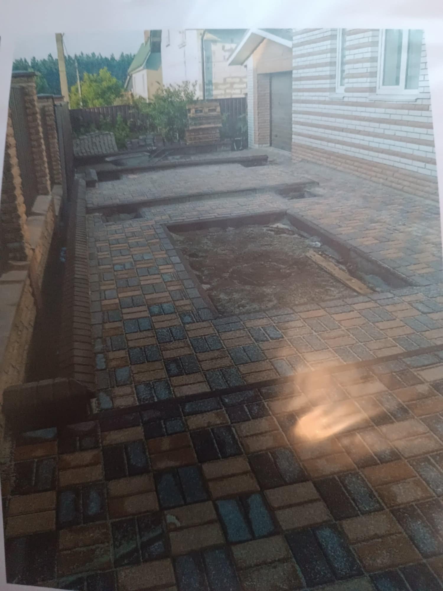 Ułożona kostka brukowa w odcieniach brązu i szarości tworzy wzór na podjeździe. Fragmenty kostki brakują, ukazując przygotowane podłoże. Widoczny mur z cegły i budynek.