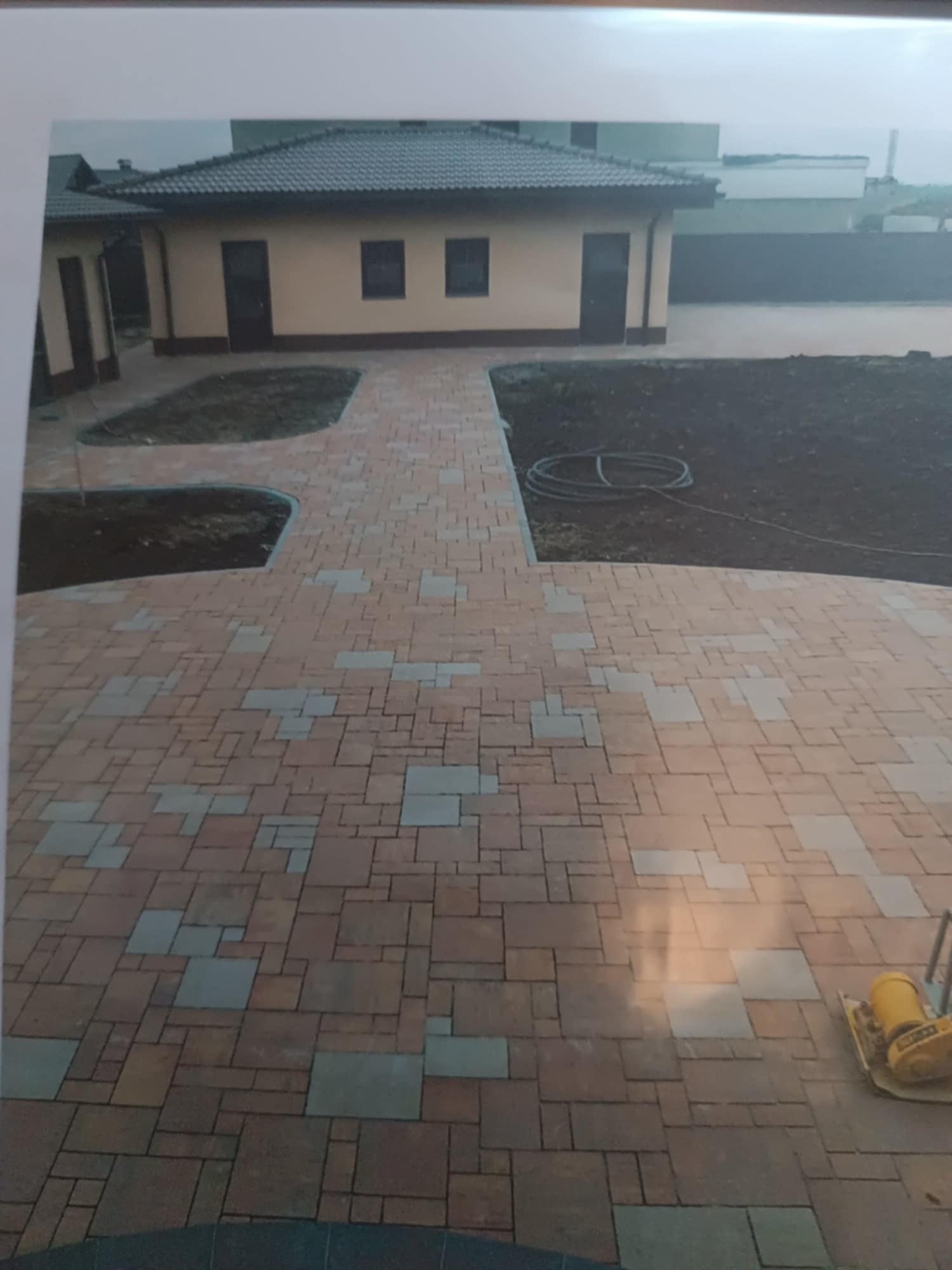 Ułożona kostka brukowa w odcieniach brązu i szarości prowadzi do budynku z ciemnym dachem. W tle widać fragment krajobrazu i żółtą zagęszczarkę.