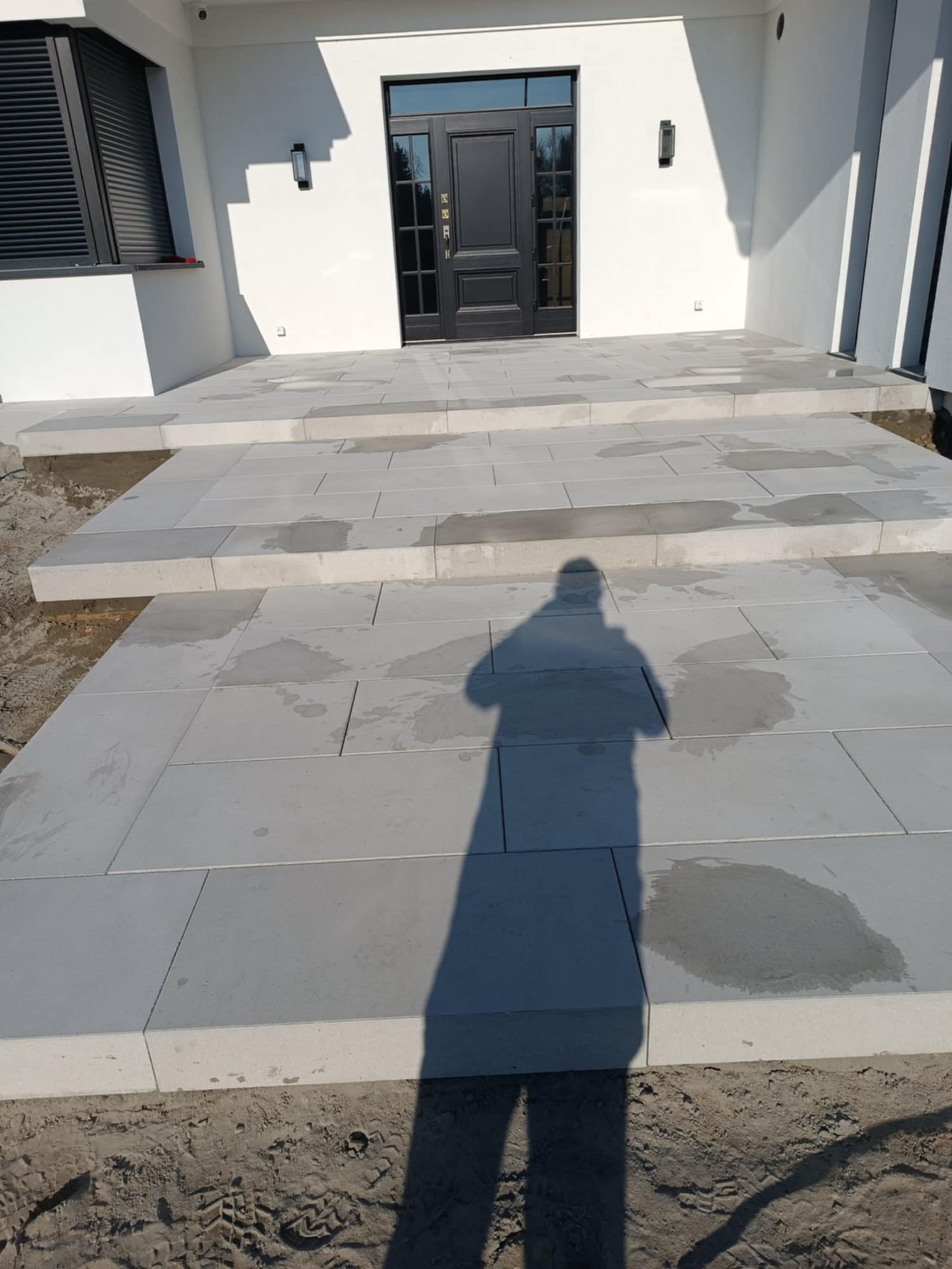 Nowoczesne schody wejściowe z jasnoszarej kostki brukowej, prowadzące do czarnych drzwi. Widoczny cień osoby fotografującej. Minimalistyczny design.